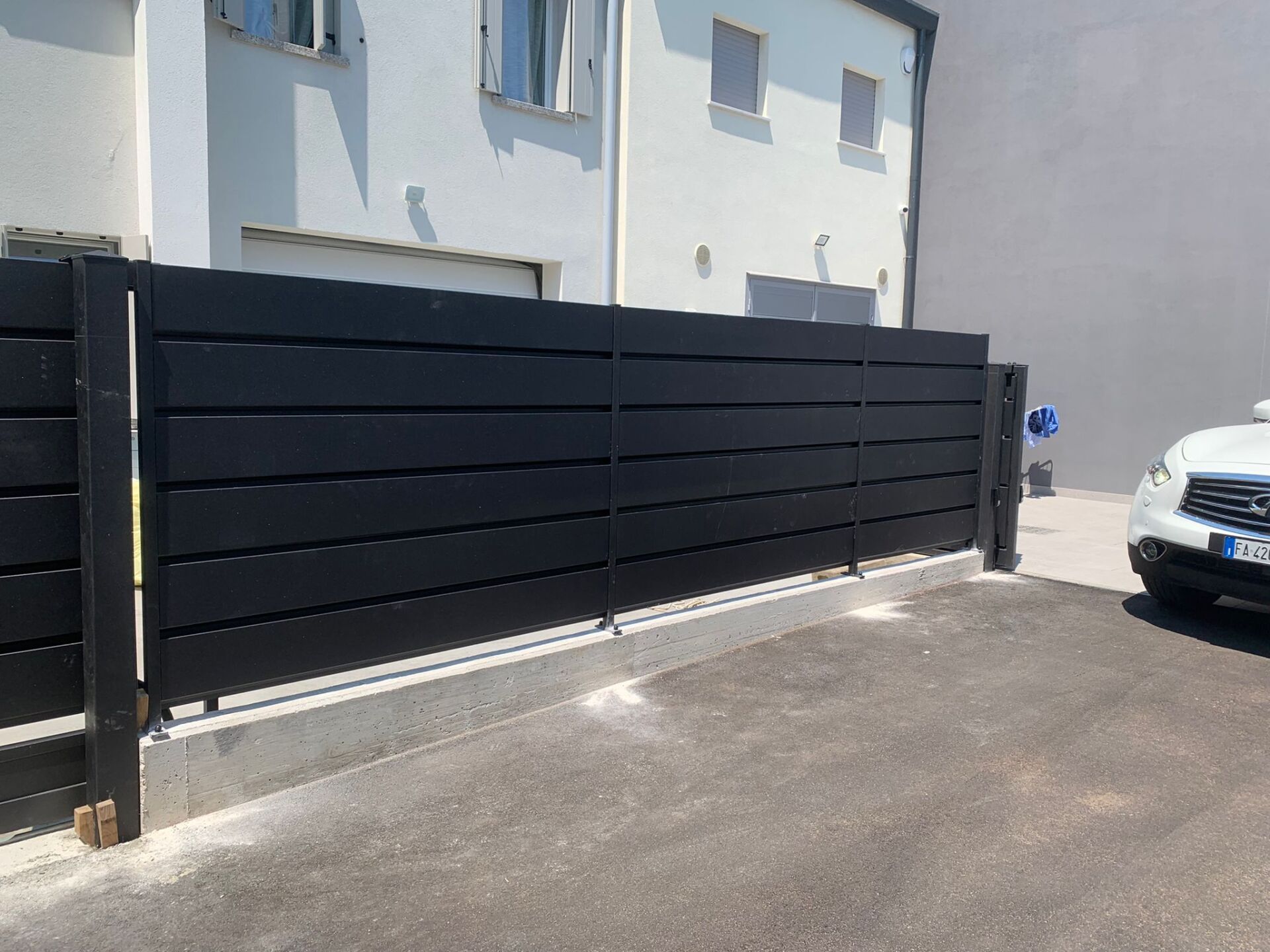 Recinzione nera a doghe orizzontali davanti a un edificio bianco e a un vialetto d'accesso. Un'auto bianca è sulla destra.