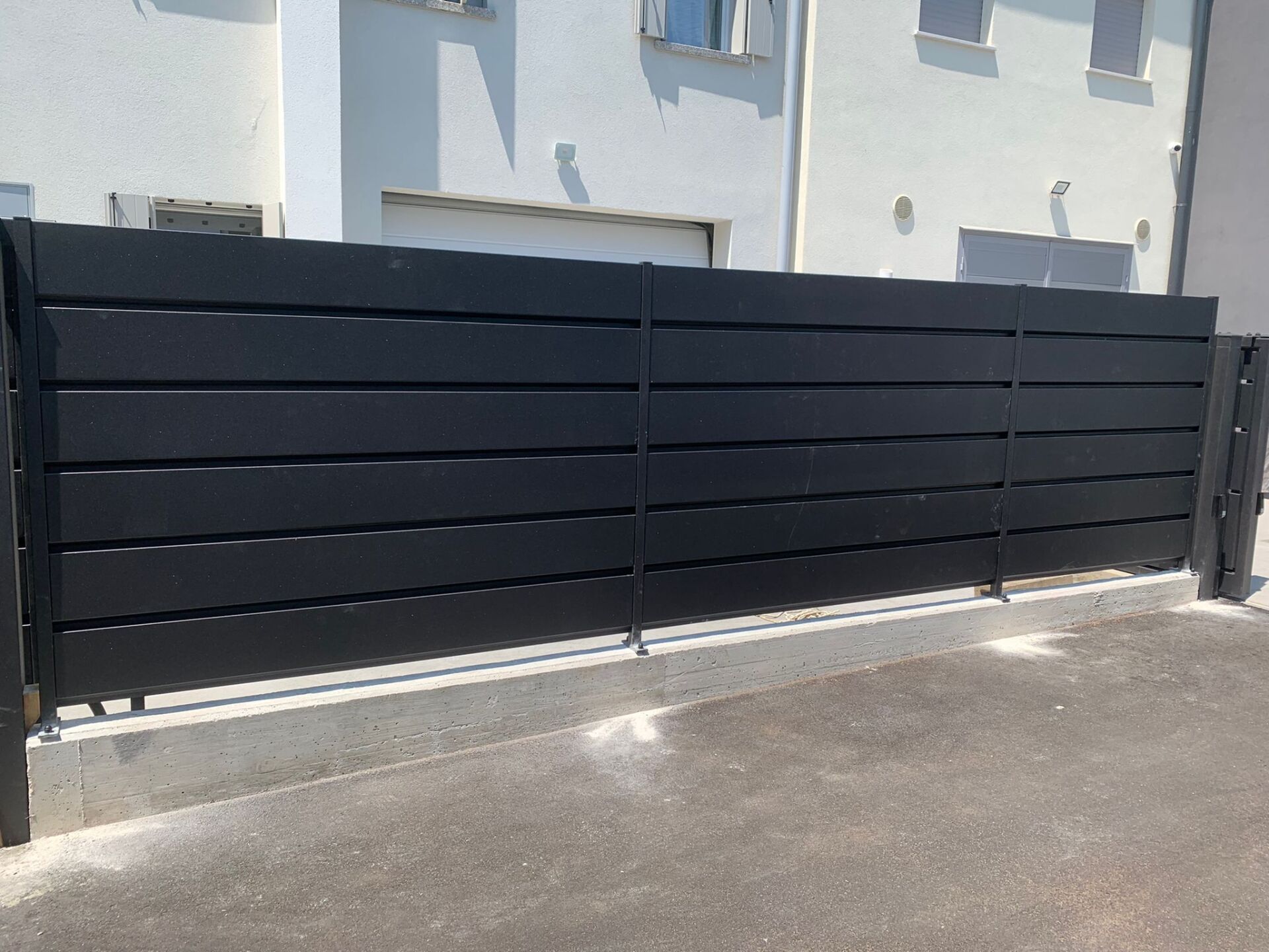 Recinzione nera con listelli orizzontali davanti a un edificio di colore chiaro.