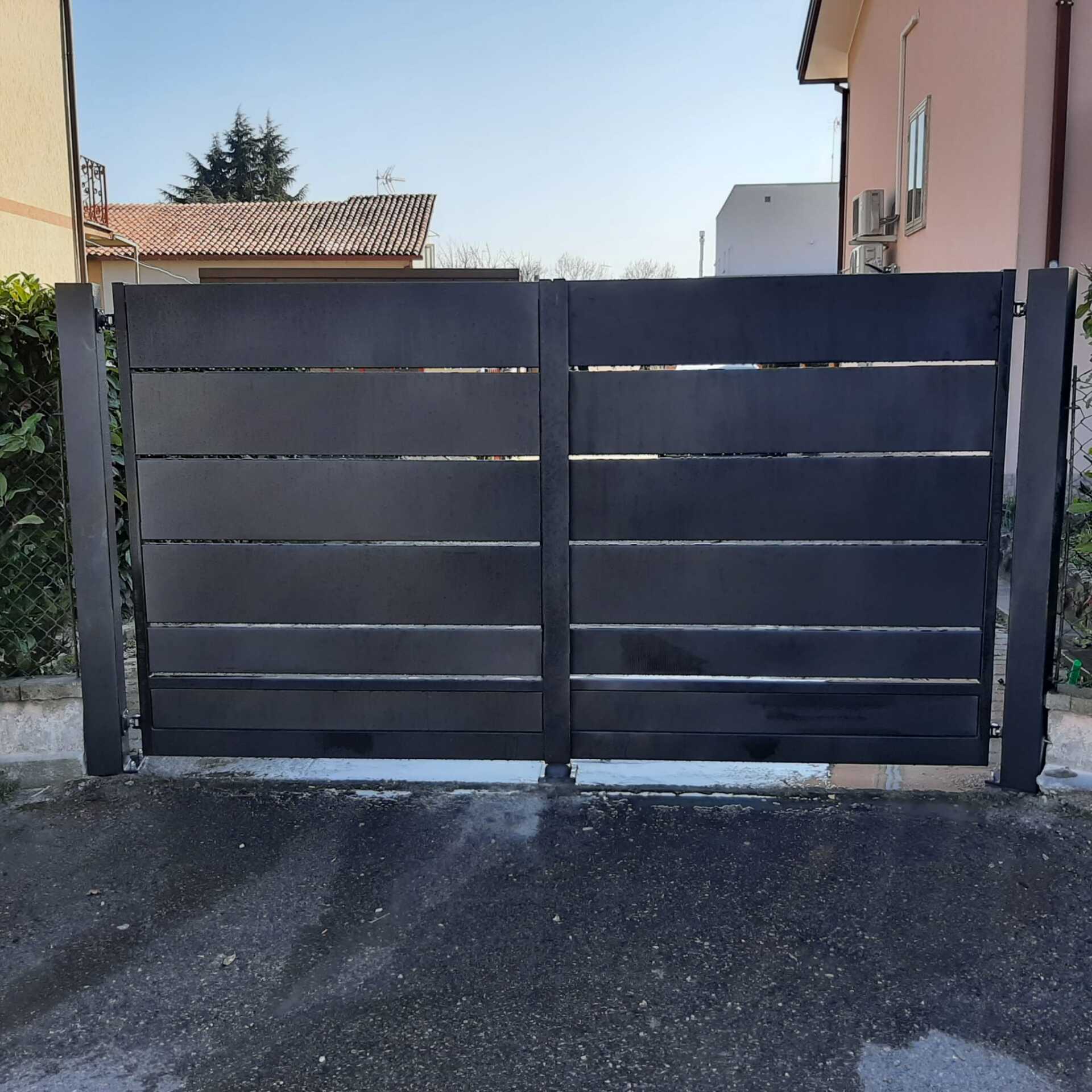 Cancello nero a stecche orizzontali, chiuso, davanti a una casa. Vialetto asfaltato, cielo azzurro.