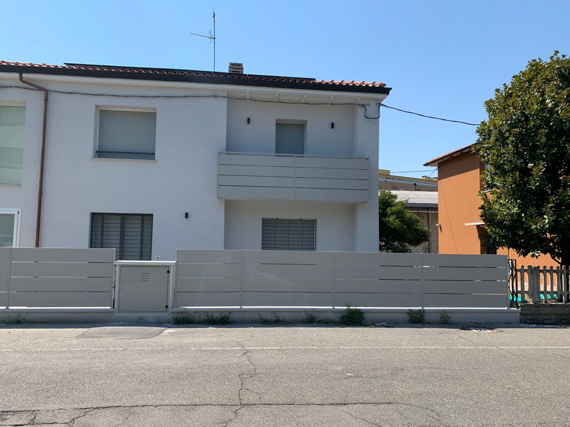 Casa bianca a due piani con recinzione grigia e balcone in una giornata di sole.