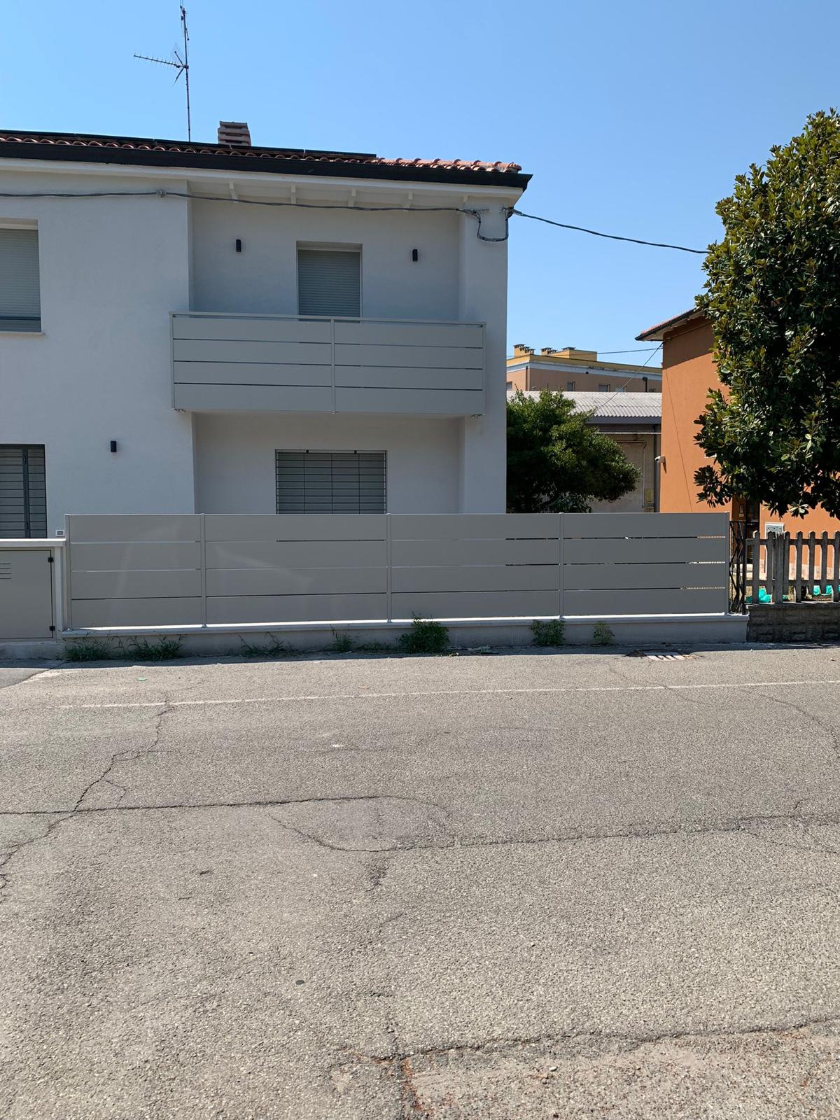 Edificio bianco a due piani con recinzione e balcone grigio chiaro. Cielo azzurro, strada asfaltata e albero.