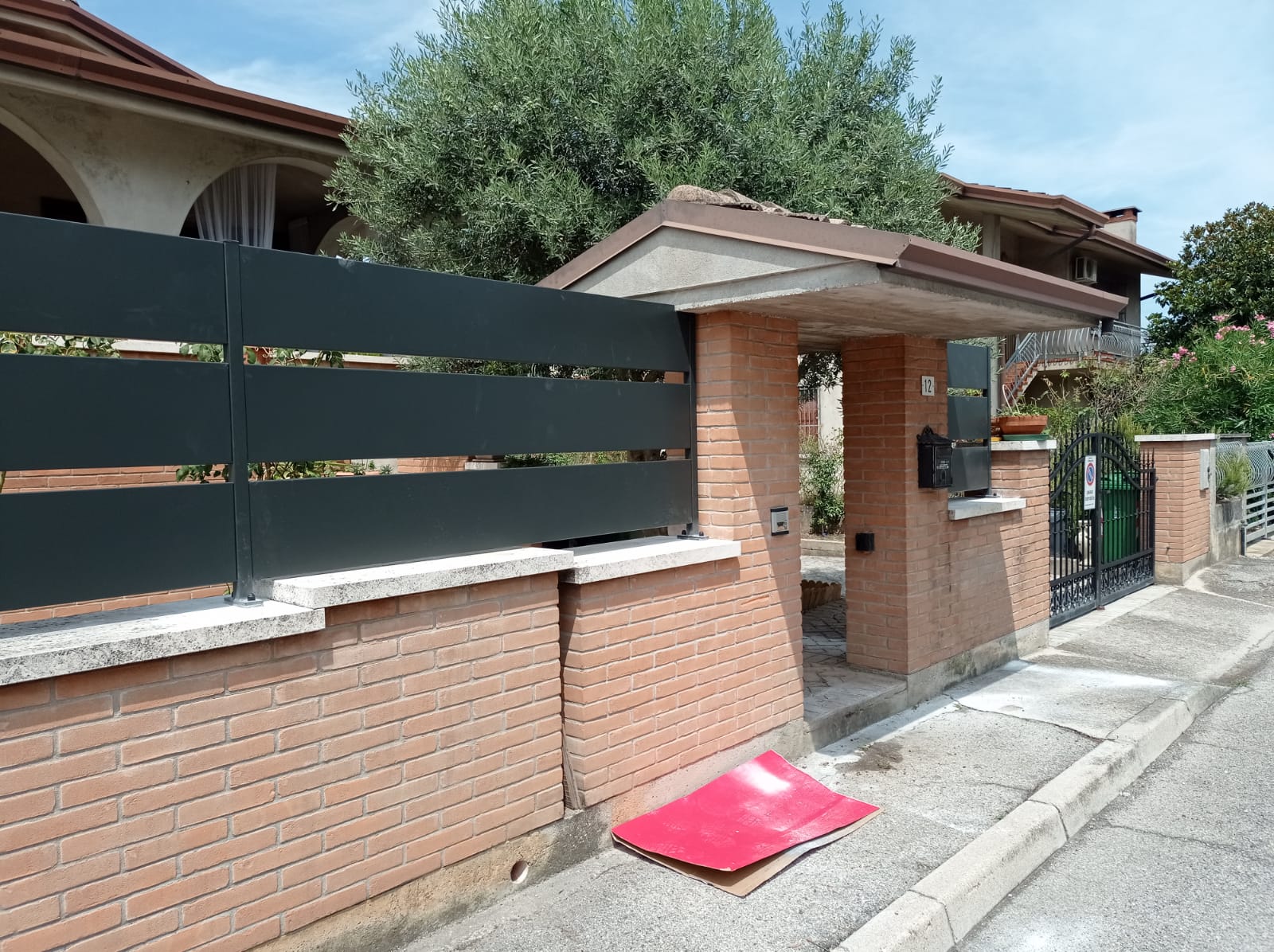 Muro di mattoni e recinzione in metallo nero con cancello d'ingresso, cassetta della posta e cartone rosso sul marciapiede.