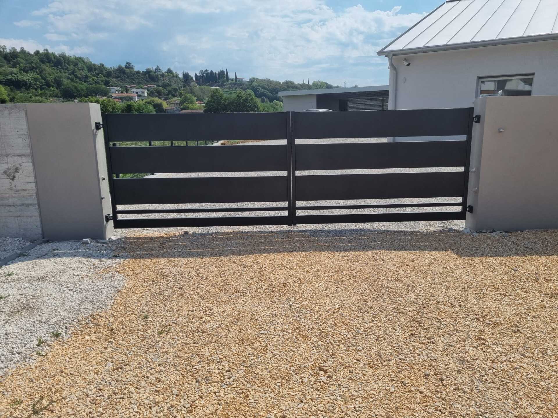 Cancello in metallo nero con stecche orizzontali, vialetto in ghiaia, muri grigi, edificio bianco.