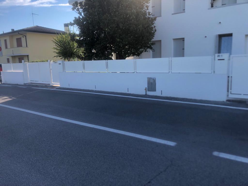Muro di recinzione bianco lungo una strada di fronte a un edificio bianco in una giornata di sole.