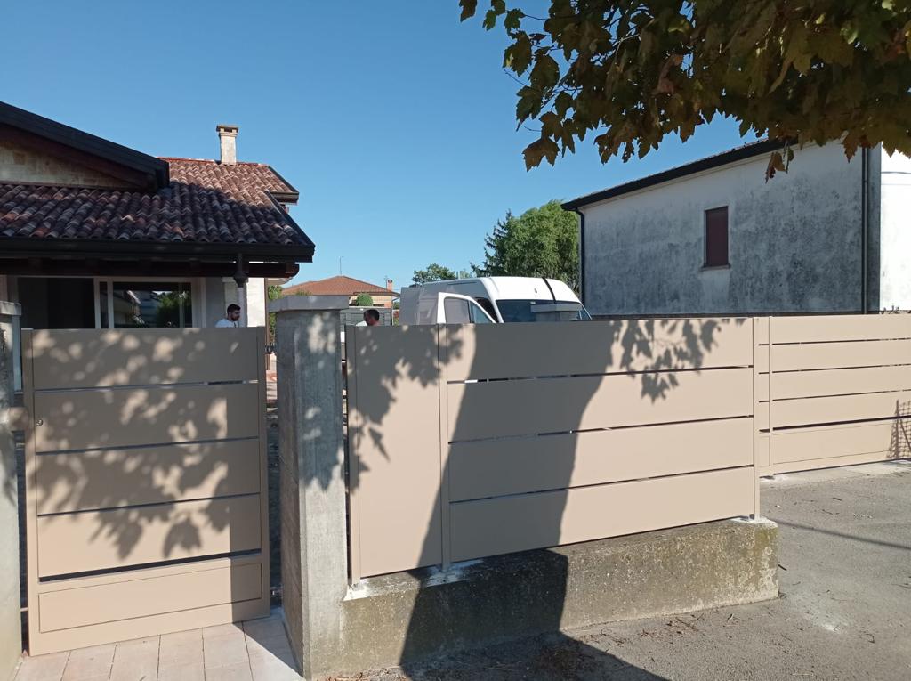 Recinzione e cancello con listelli orizzontali beige davanti a una casa, con cielo azzurro e rami di alberi sopra di noi.