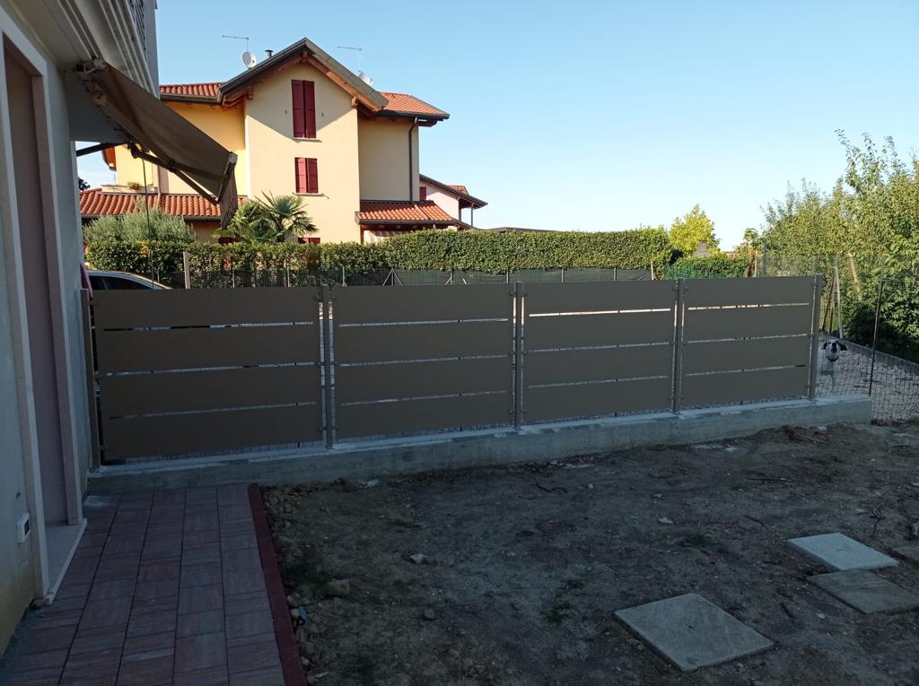 Recinzione in listelli orizzontali marroni con pali argentati e una casa sullo sfondo.