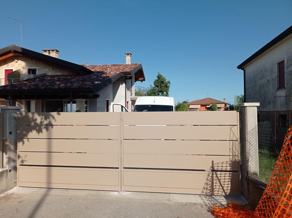 Cancello marrone chiaro con stecche orizzontali chiuso davanti a un vialetto, case sullo sfondo, cielo azzurro.