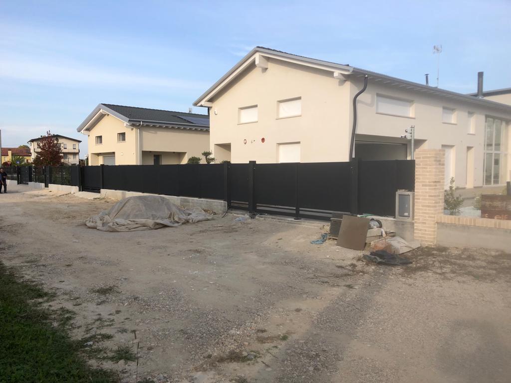 Recinzione nera davanti a due case beige con le persiane chiuse. Vialetto di ghiaia, cielo nuvoloso.