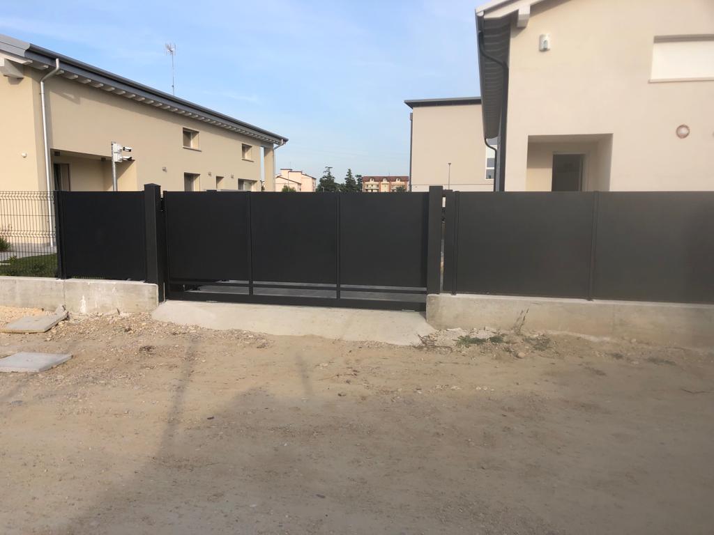 Cancello e recinzione in metallo nero davanti a case beige in una giornata di sole, con un vialetto di ghiaia.