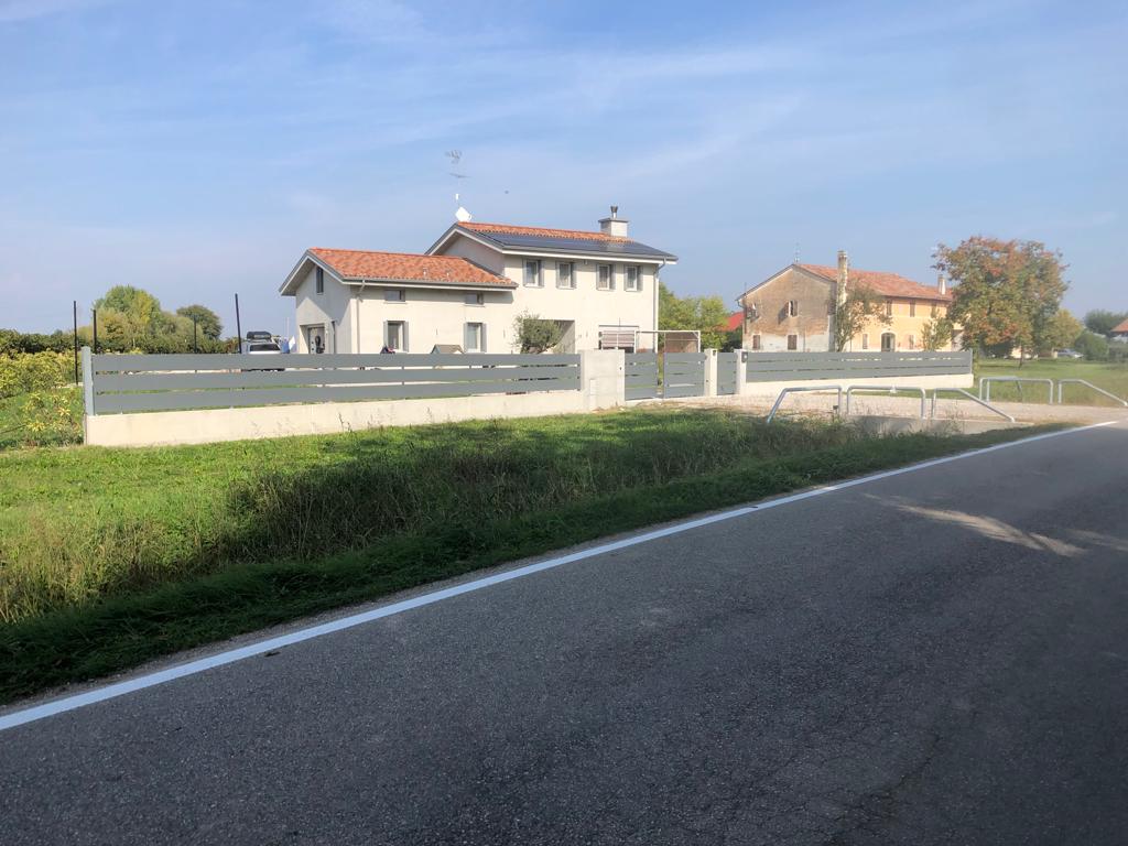 Una casa con una recinzione grigia e un cancello su un prato verde accanto a una strada, sotto un cielo azzurro.