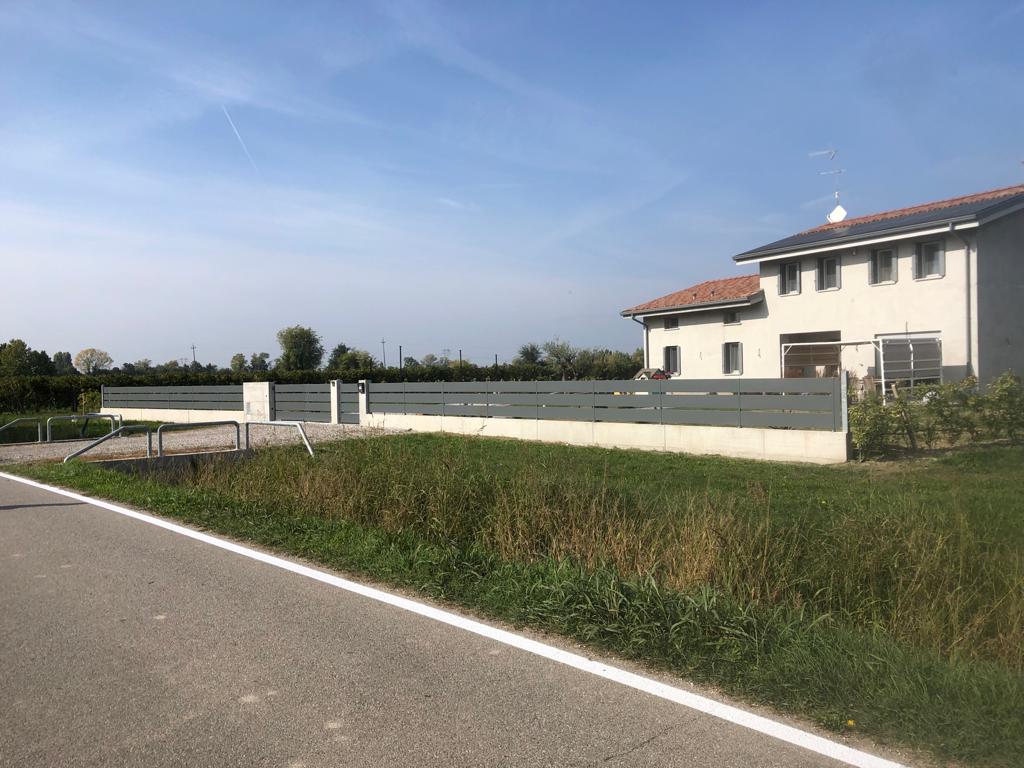 Casa con recinzione e strada grigie, campo verde, cielo azzurro.