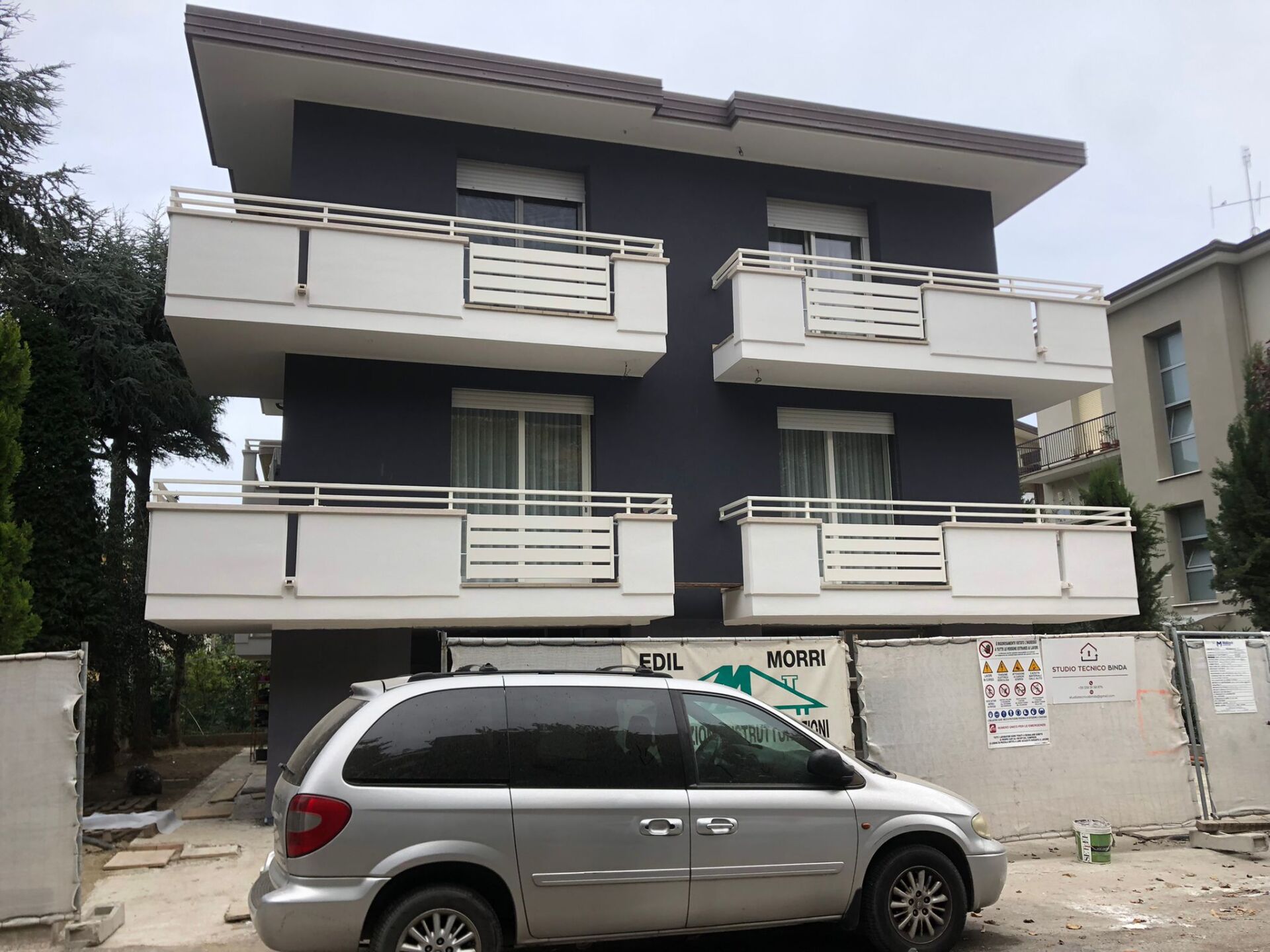 Edificio di tre piani con balconi bianchi ed esterni grigi. Un minivan argentato parcheggiato di fronte.