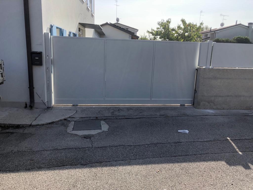 Cancello scorrevole grigio chiuso davanti a una casa. Strada asfaltata e blocchi di cemento.