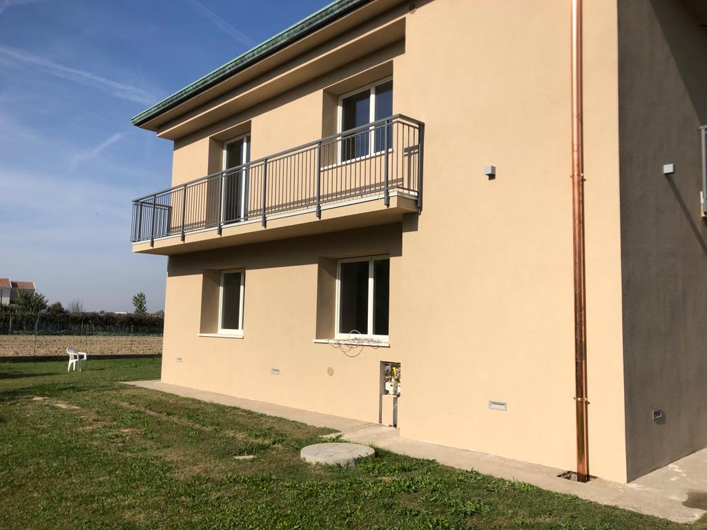 Casa color marrone chiaro a due piani con balcone e grondaia in rame, circondata da erba verde.