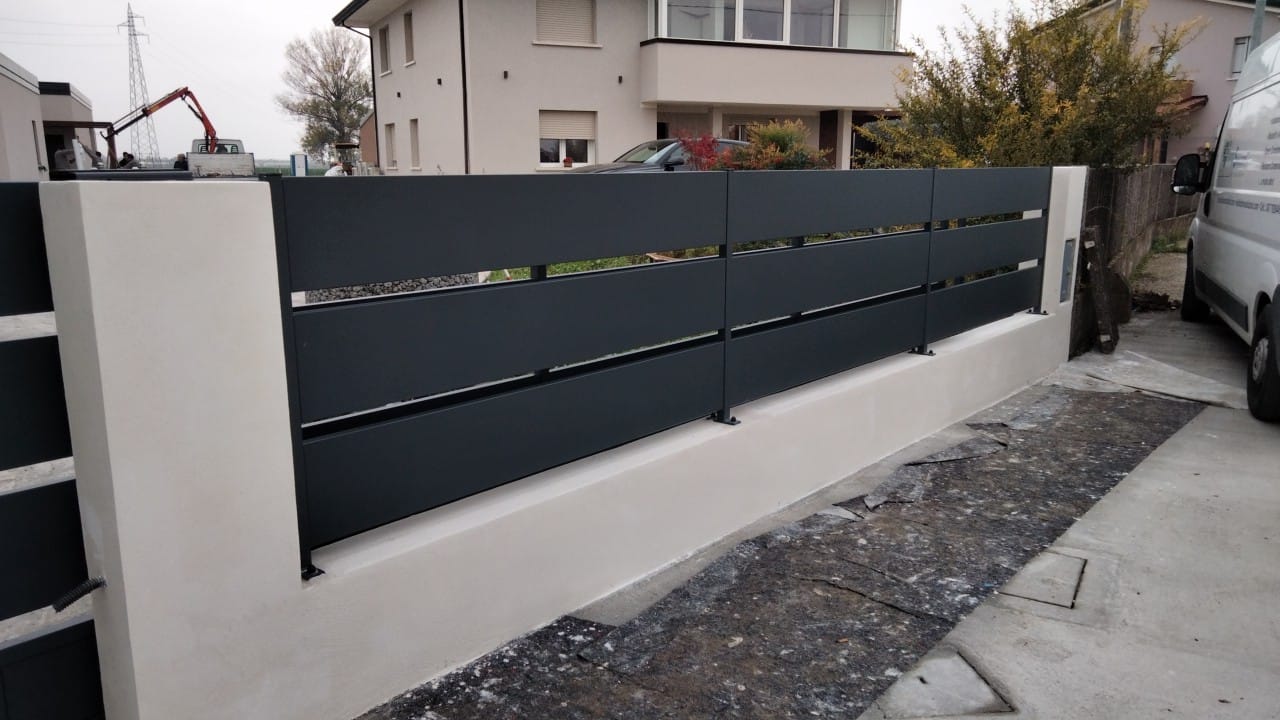 Recinzione grigia con listelli orizzontali sopra un muro di cemento bianco, posta di fronte a una casa.