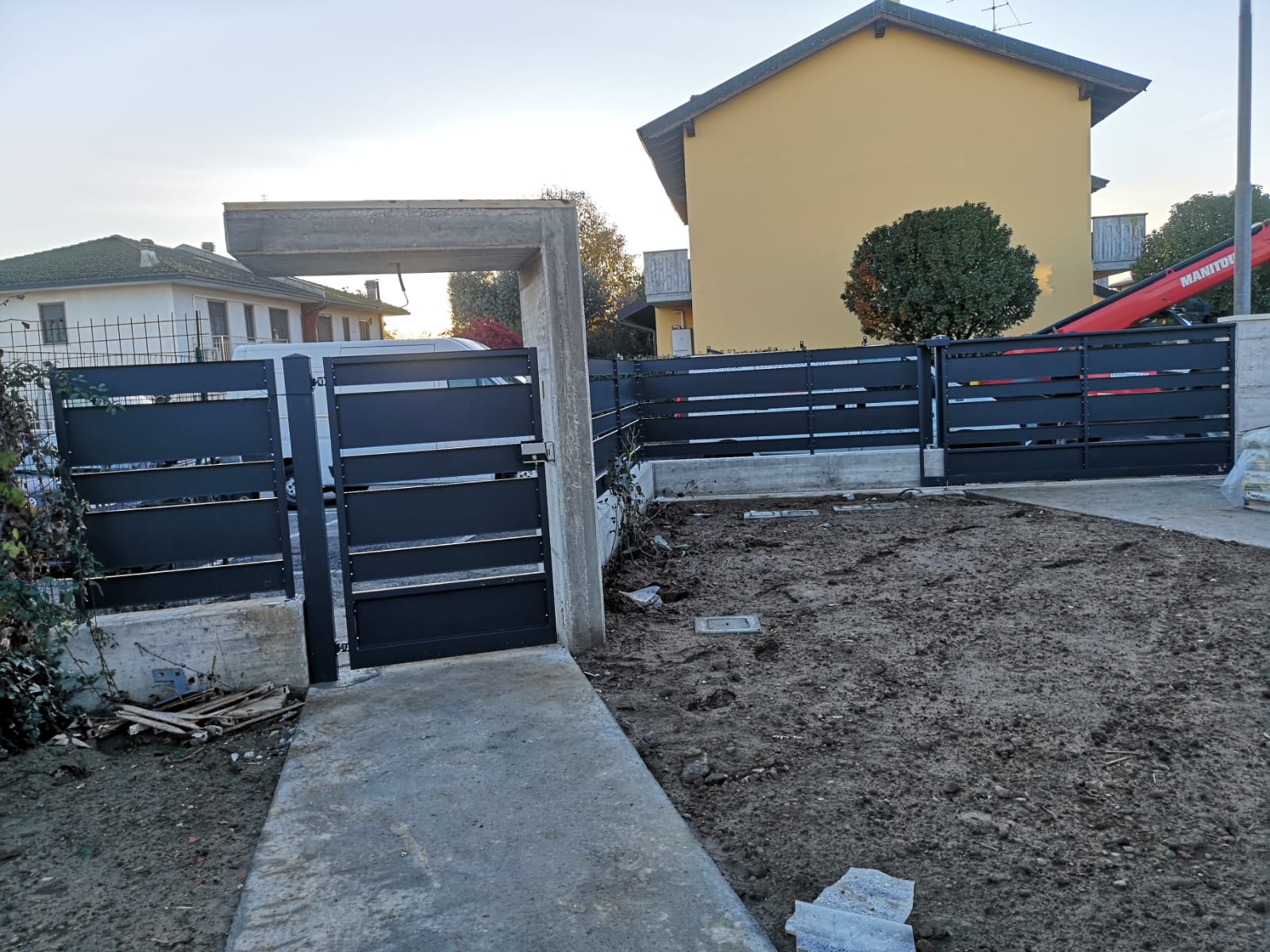 Cancello e recinzione in metallo nero davanti a una casa, con un cortile sterrato in primo piano.