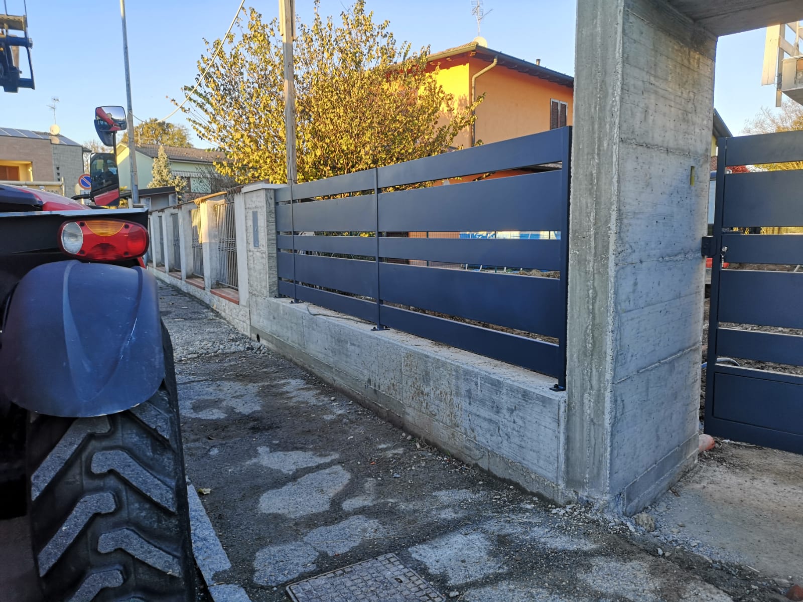 Recinzione nera con stecche orizzontali costruita su una fondazione in cemento; in primo piano c'è un trattore.