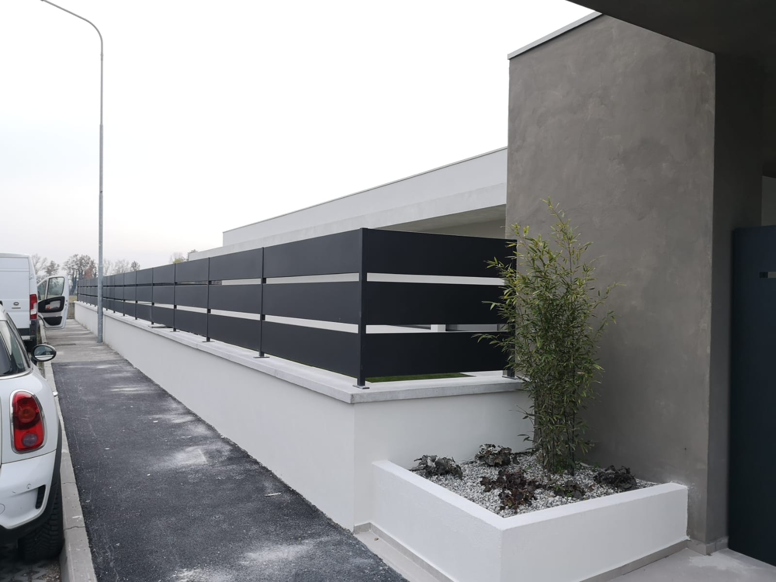 Recinzione a doghe orizzontali bianche e nere in cima a un muro bianco. Dietro la recinzione si trova un edificio moderno.