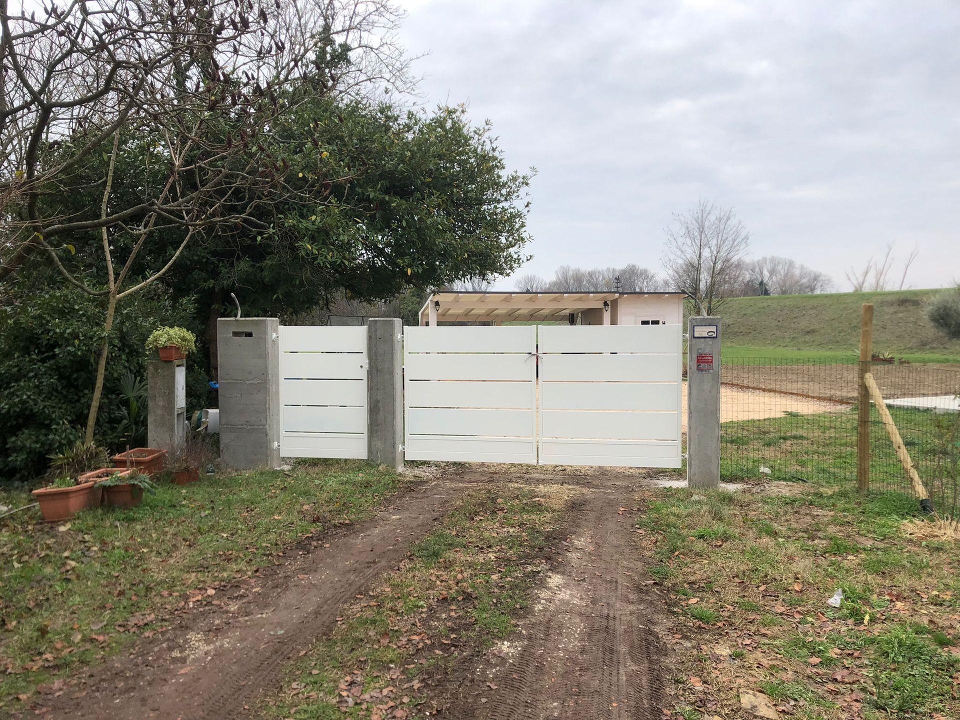 Cancello d'ingresso bianco di una casa, con vialetto sterrato e verde circostante in una giornata nuvolosa.
