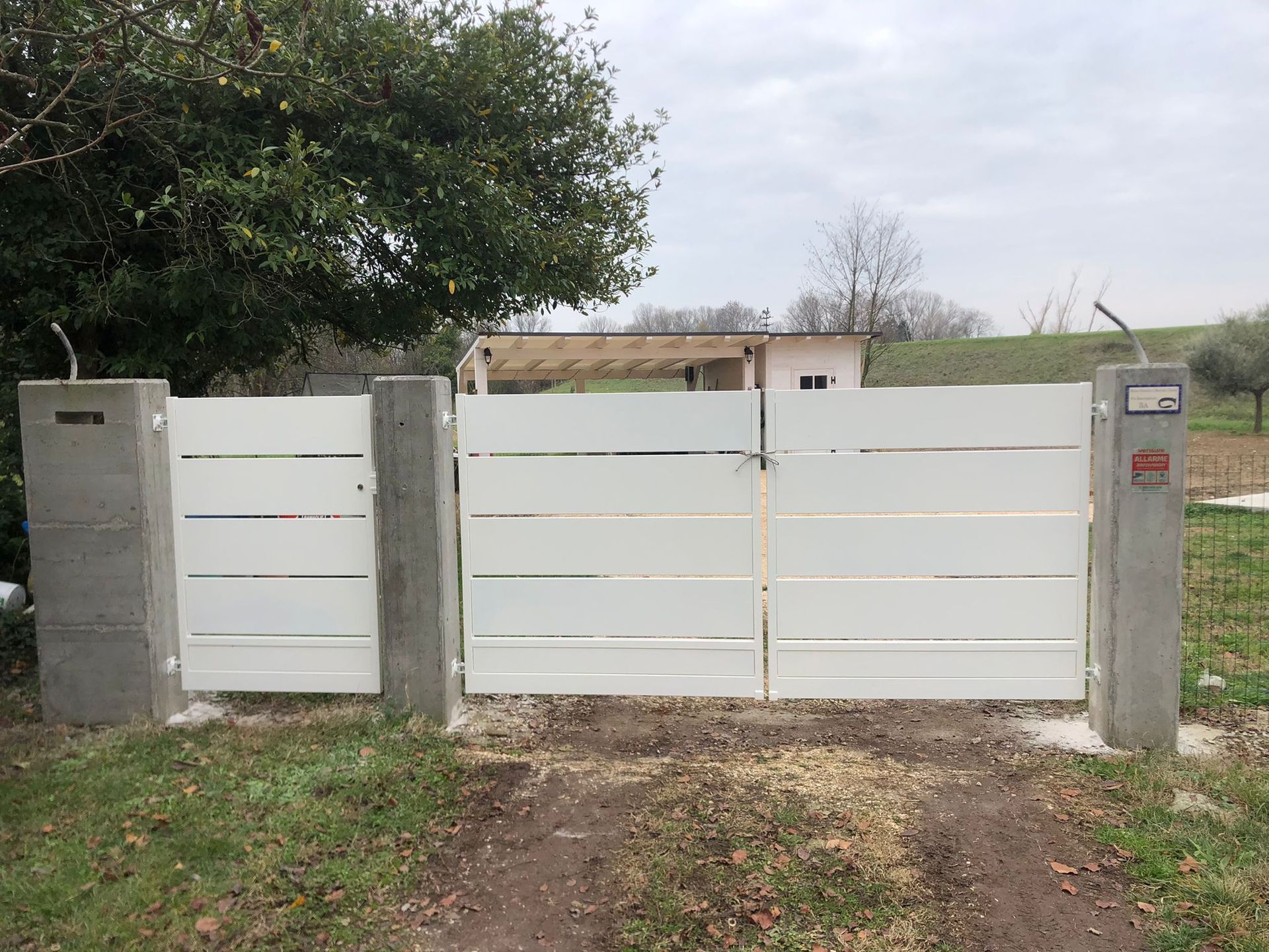 Cancello e pali di recinzione in metallo bianco con vialetto d'accesso, sullo sfondo di un cielo nuvoloso.