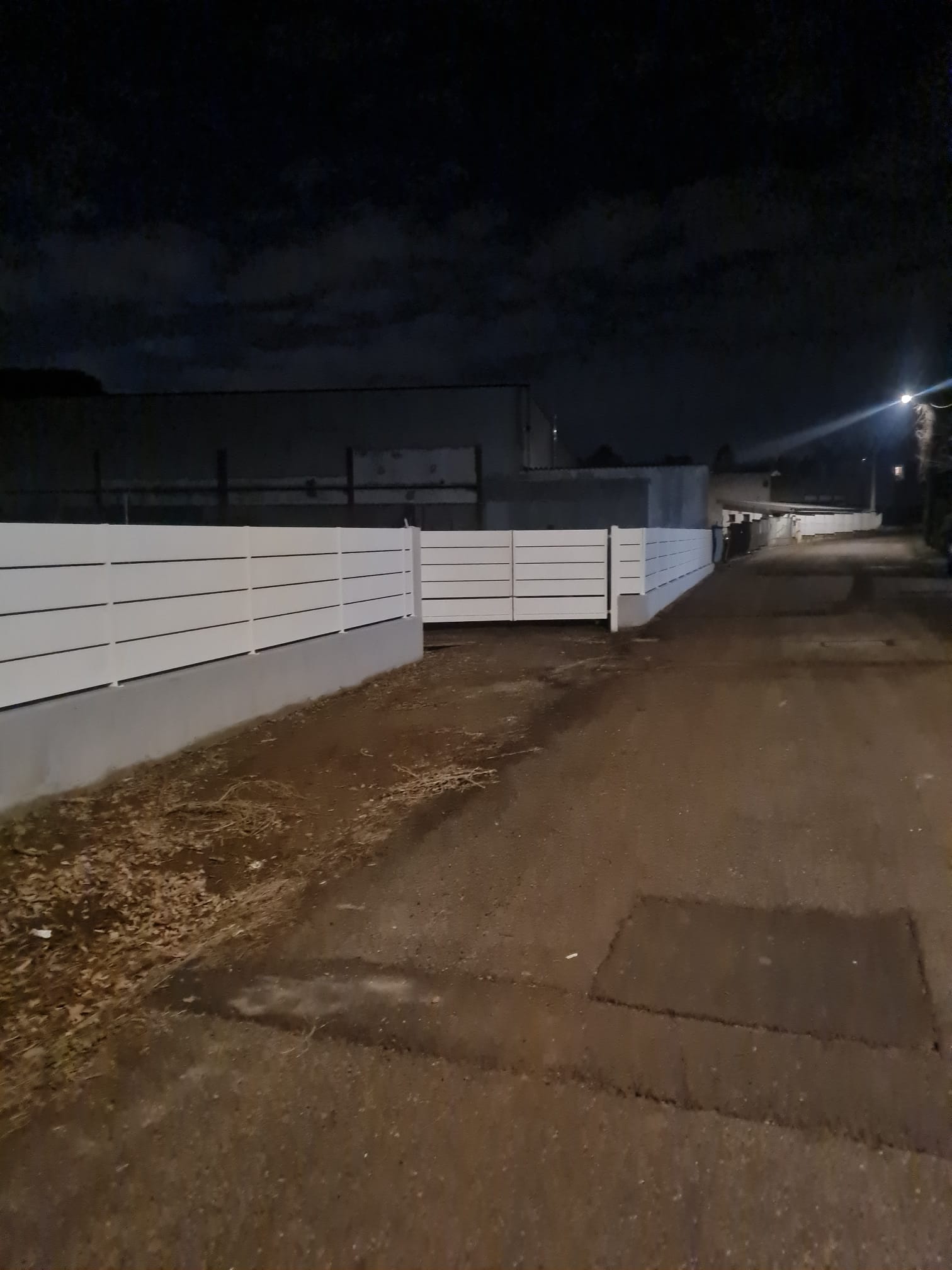 Vicolo buio di notte con recinzione bianca a listelli orizzontali, strada asfaltata ed edifici sullo sfondo.