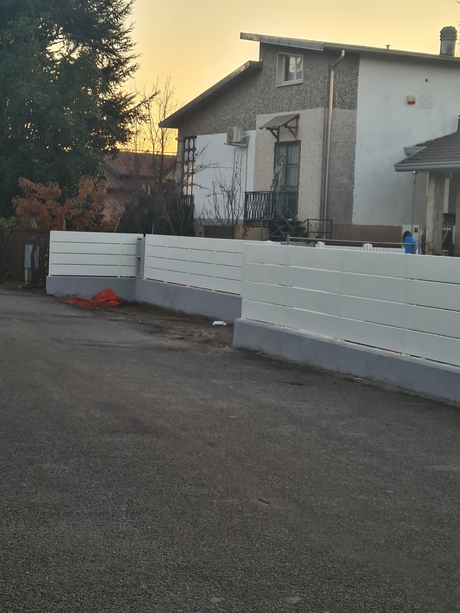 Una staccionata bianca lungo una strada sterrata che conduce verso una casa. Il cielo è di una tonalità arancione.