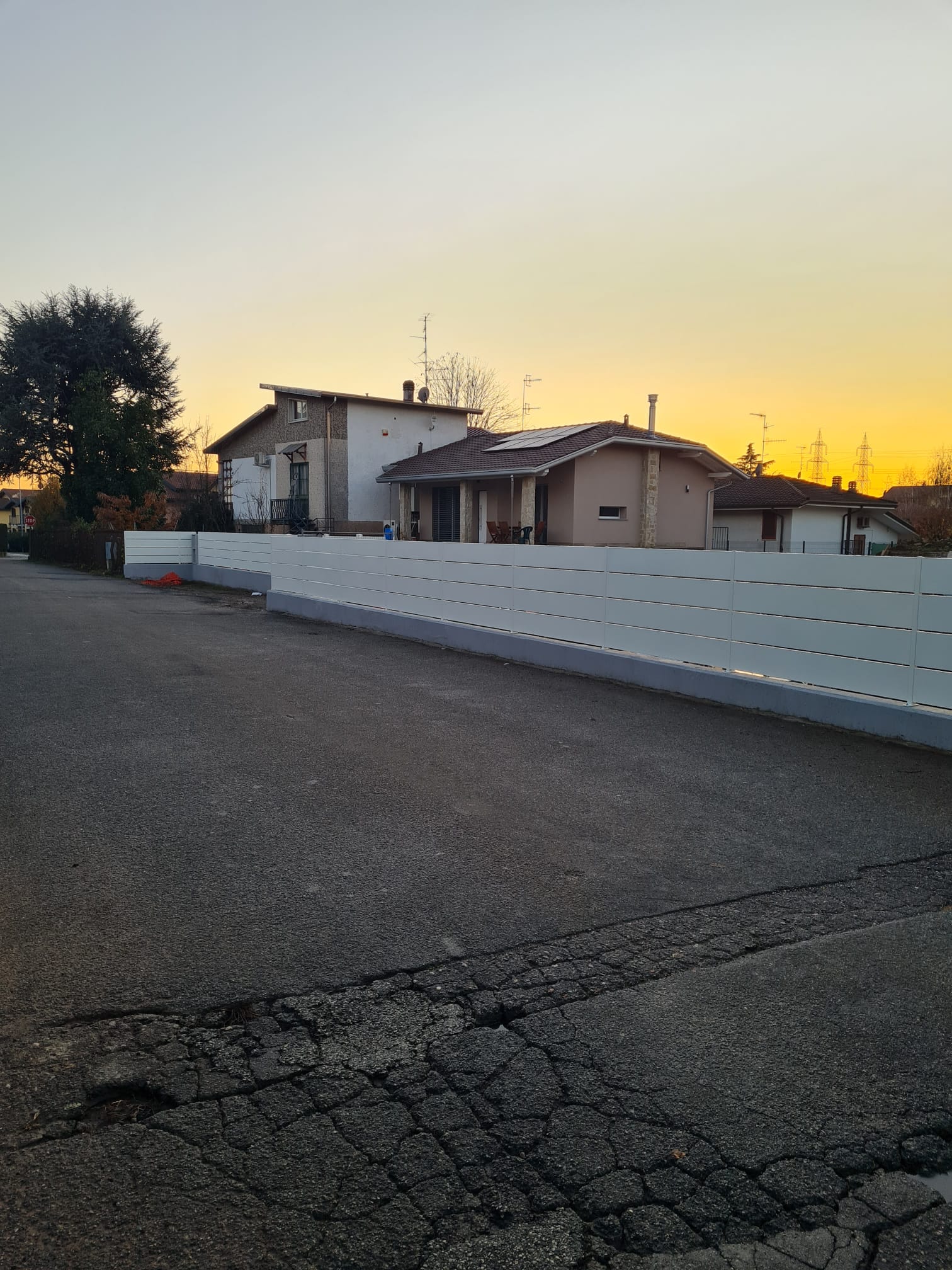 Strada con pavimentazione crepata, una lunga recinzione bianca e case sullo sfondo del cielo al tramonto.