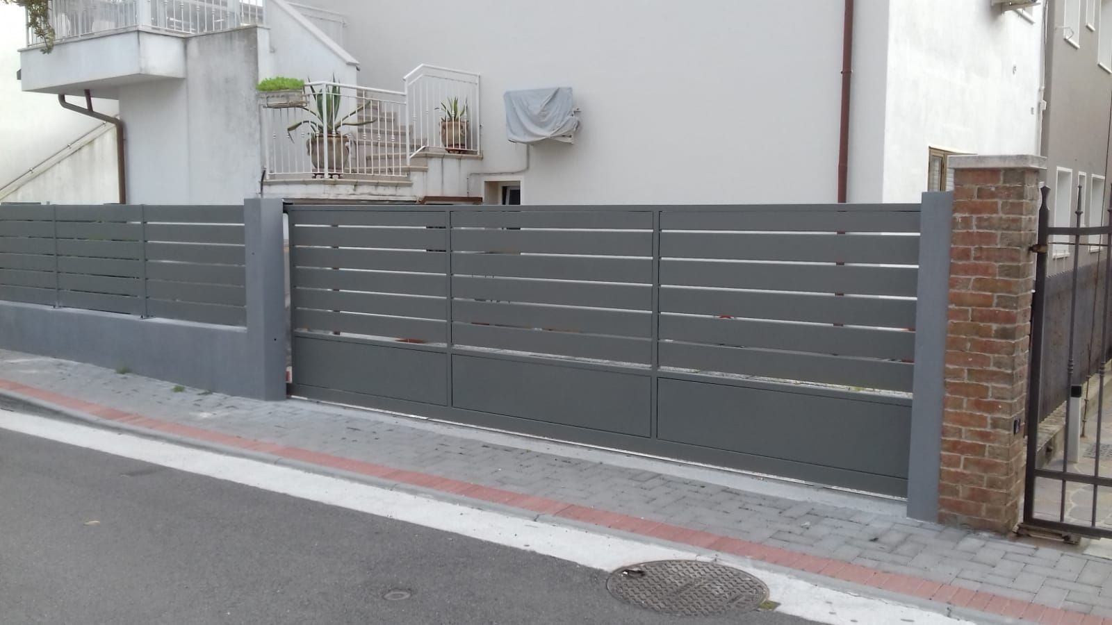 Recinzione in metallo grigio con doghe orizzontali e pilastri in mattoni lungo una strada.