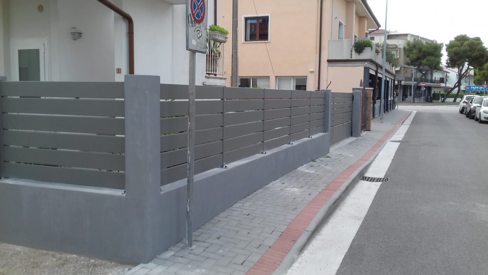 Recinzione orizzontale grigia di fronte agli edifici su una strada con auto parcheggiate.