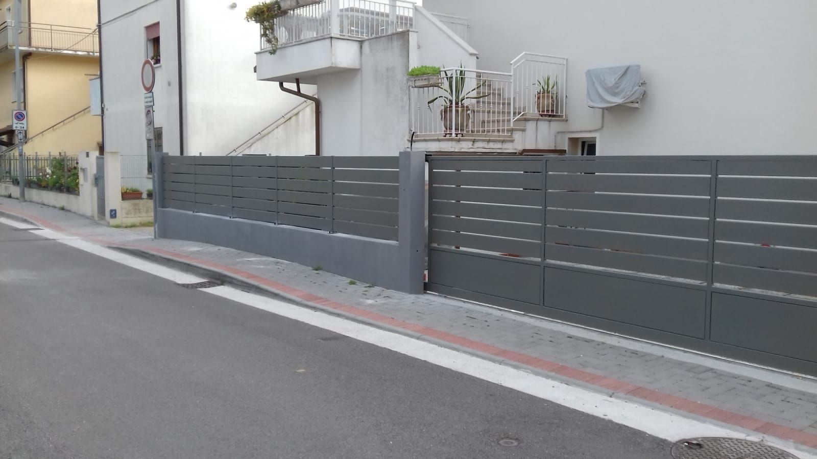 Recinzione grigia con listelli orizzontali lungo una strada, di fronte a un edificio.