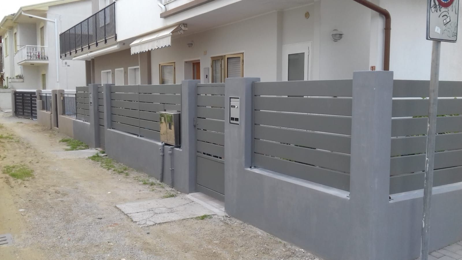 Recinzione e cancello grigi di fronte a un edificio bianco su una strada sterrata.