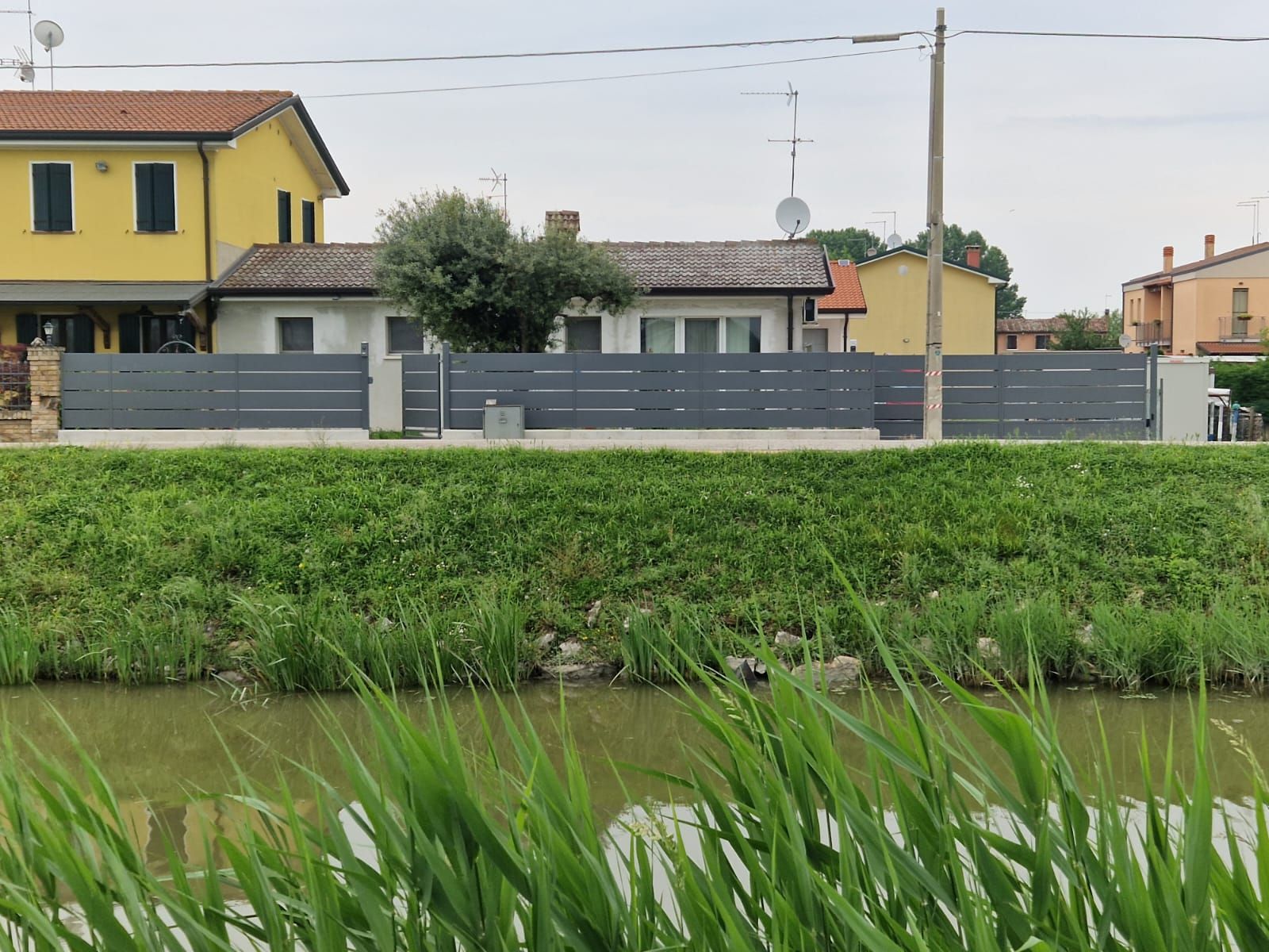 Una recinzione grigio scuro racchiude una casa dietro un argine erboso e uno specchio d'acqua, in una giornata nuvolosa.