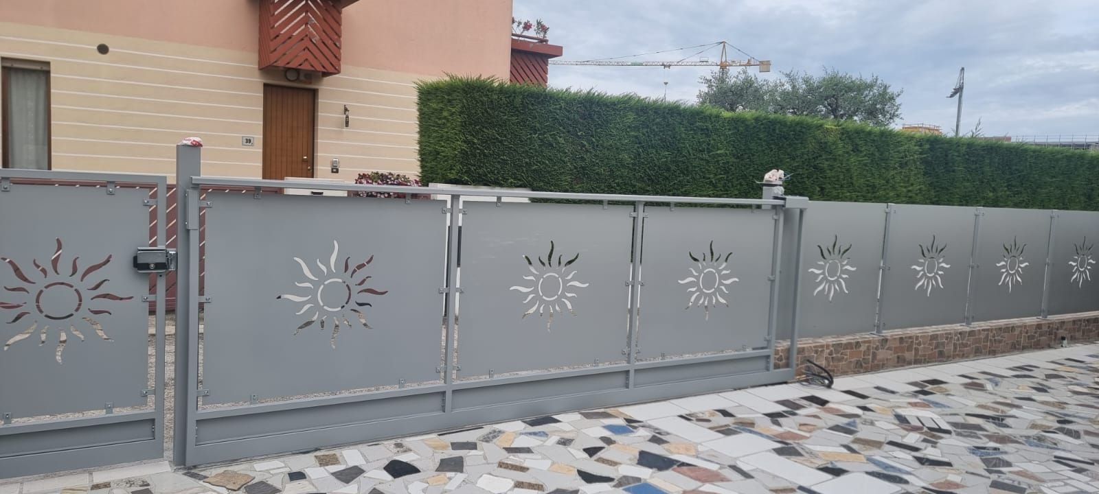 Cancello in metallo grigio con ritagli a forma di sole, una casa e una siepe verde sullo sfondo.