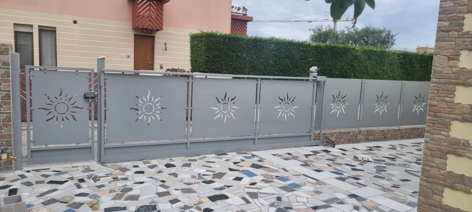 Cancello in metallo grigio con ritagli a raggiera, di fronte a una casa con vialetto in pietra e siepe verde.
