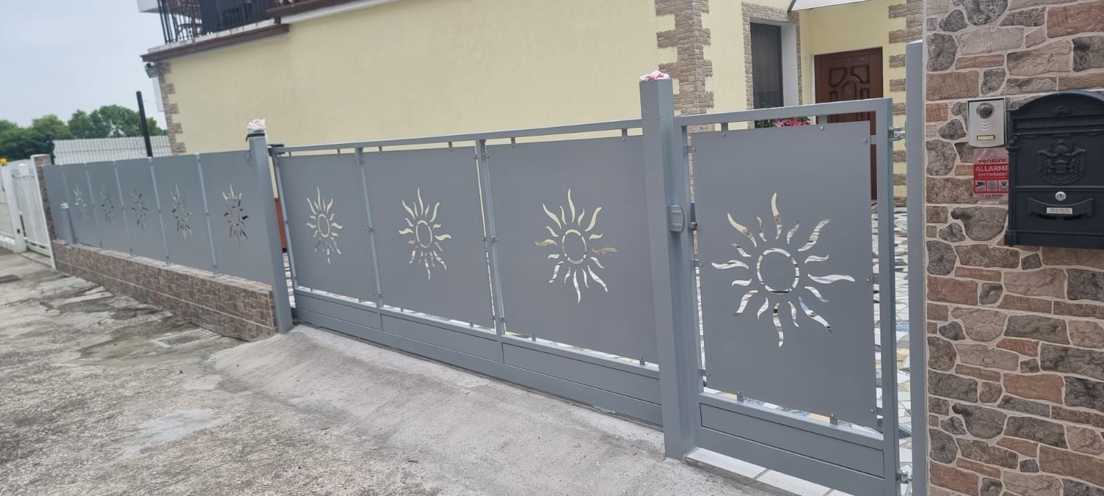 Cancello e recinzione in metallo grigio con motivi a raggi di sole davanti a una casa.