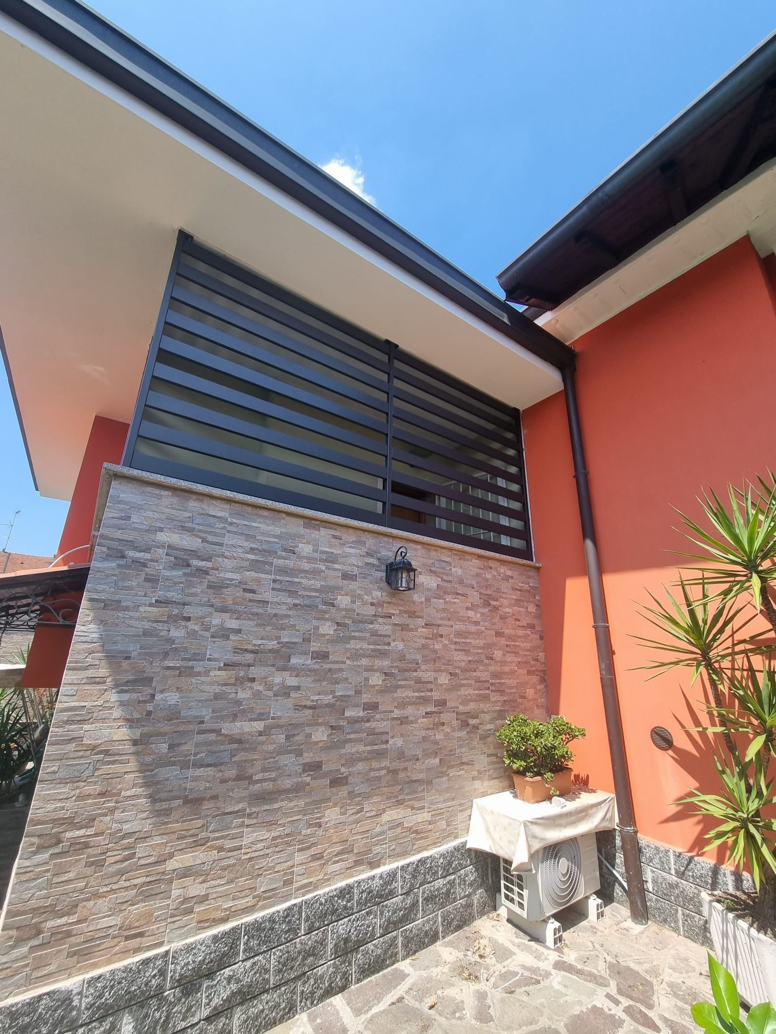 Esterno di un edificio moderno con pareti arancioni e in pietra, schermo a doghe nere e cielo azzurro.