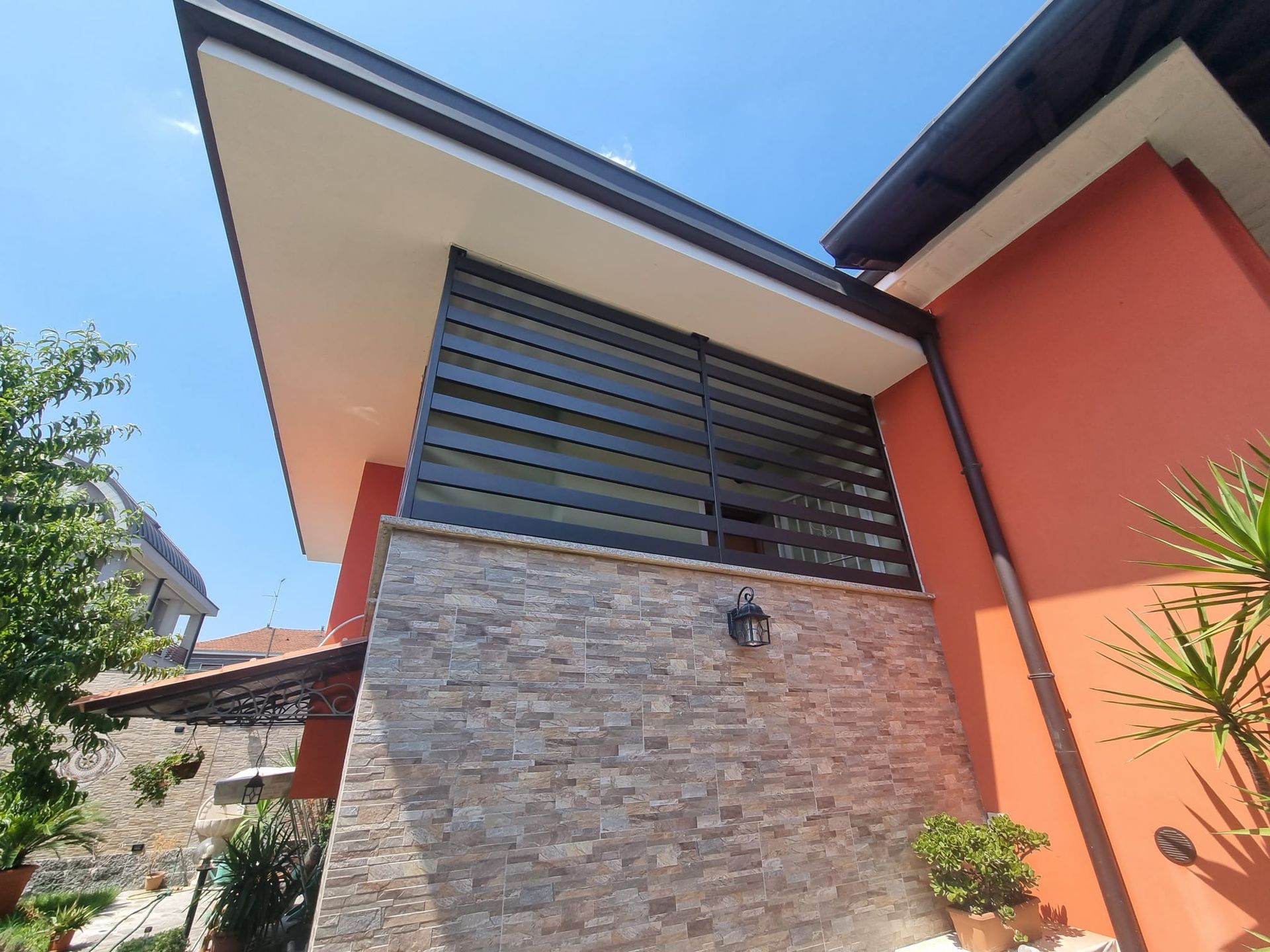 Esterno di un edificio con pareti arancioni, facciata in pietra e persiane orizzontali scure sotto una sporgenza bianca.