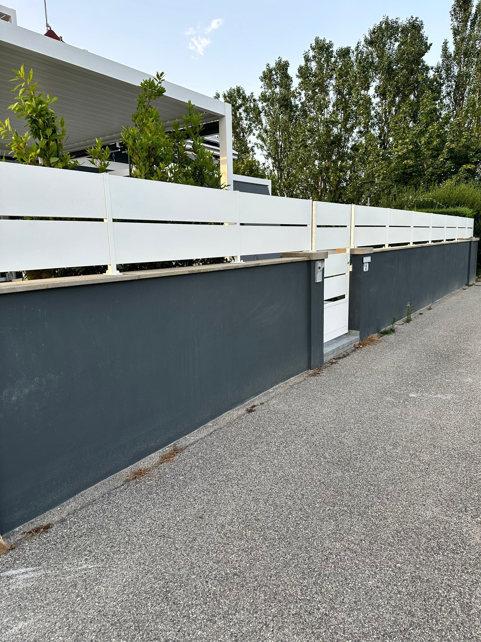 Recinzione bianca a doghe in cima a un muro grigio con un piccolo cancello bianco, alberi e cielo sullo sfondo.