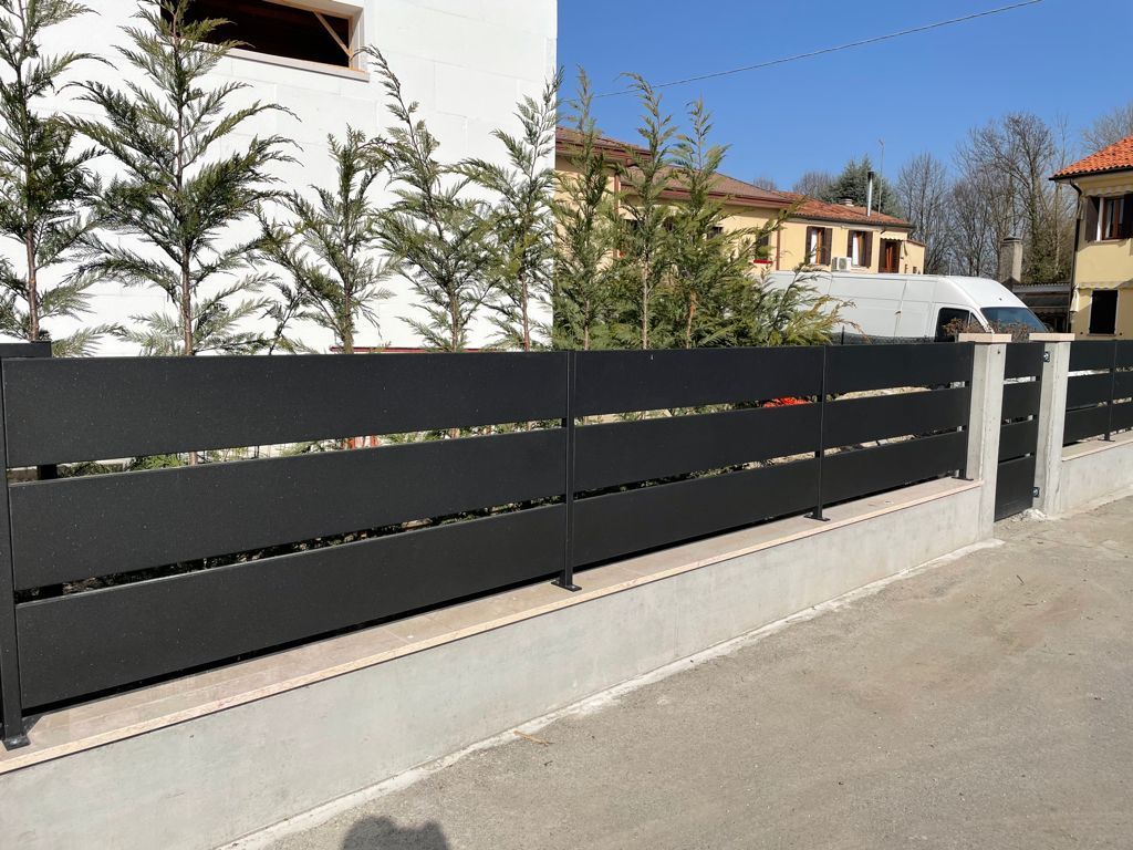 Recinzione nera con listelli orizzontali su una base di cemento, con alberi verdi ed edifici sullo sfondo.