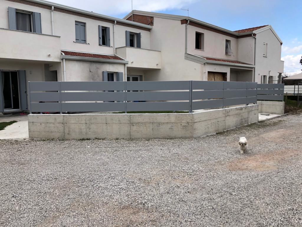 Recinzione grigia di fronte a un edificio multifamiliare di colore chiaro con ghiaia in primo piano.