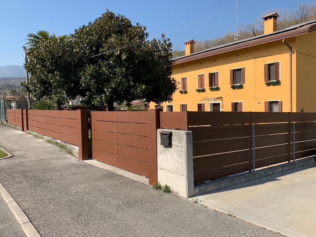 Recinzione con stecche marroni e cancello di fronte a un edificio giallo con un albero su una strada asfaltata.