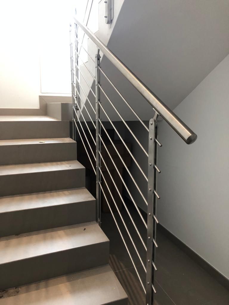 Scala con ringhiera in acciaio inox e cavi metallici. Gradini in cemento salgono, vicino a una parete bianca.