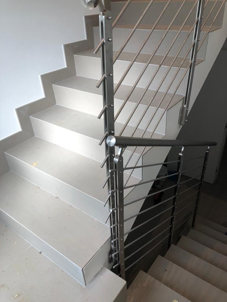 Scala con ringhiera in acciaio inox e corrimano scuro. Gradini grigi e pareti bianche.