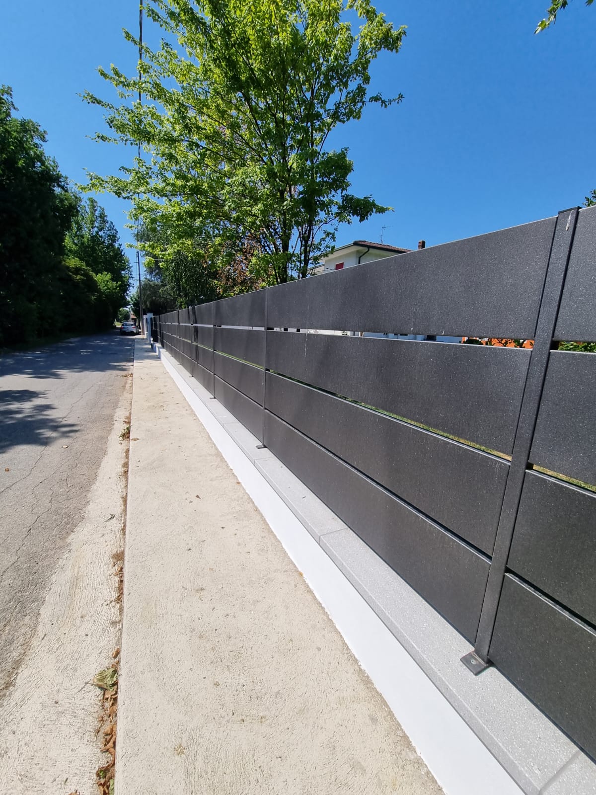 Recinzione grigia lungo un marciapiede, con un albero e una strada sullo sfondo sotto un cielo azzurro.