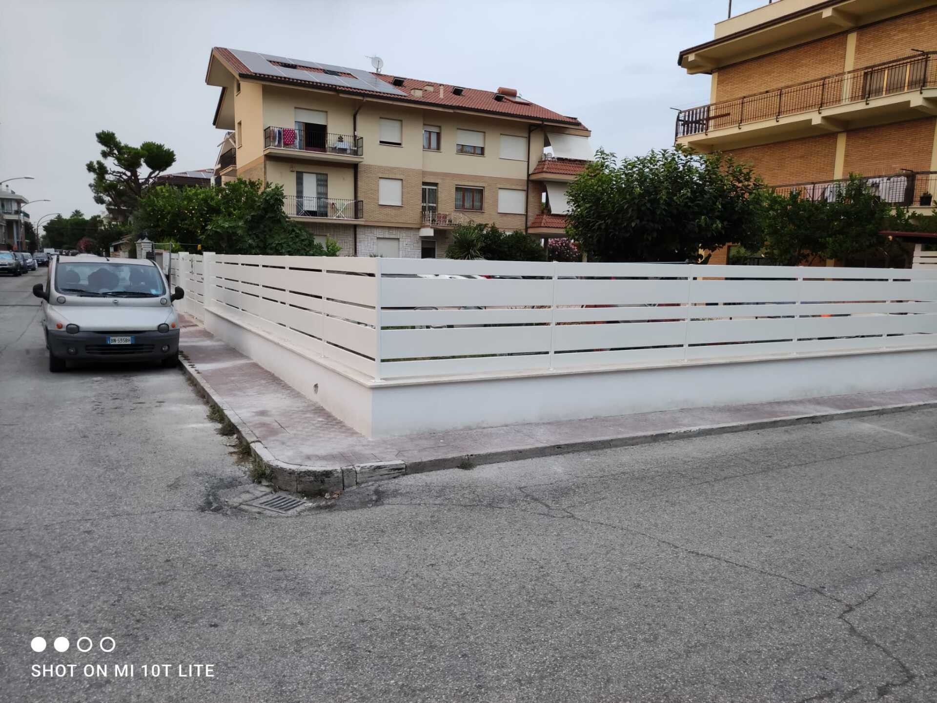 Recinzione bianca a doghe orizzontali davanti agli edifici. Un'auto è parcheggiata sulla strada.