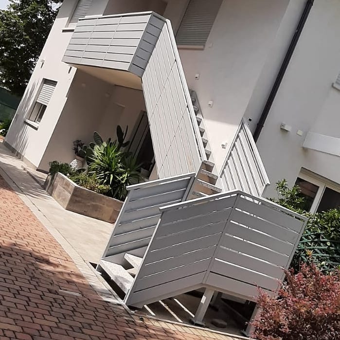 Esterno di un edificio con scale, ringhiera e finiture bianche. Vialetto pavimentato. Piante in una fioriera e cespugli.