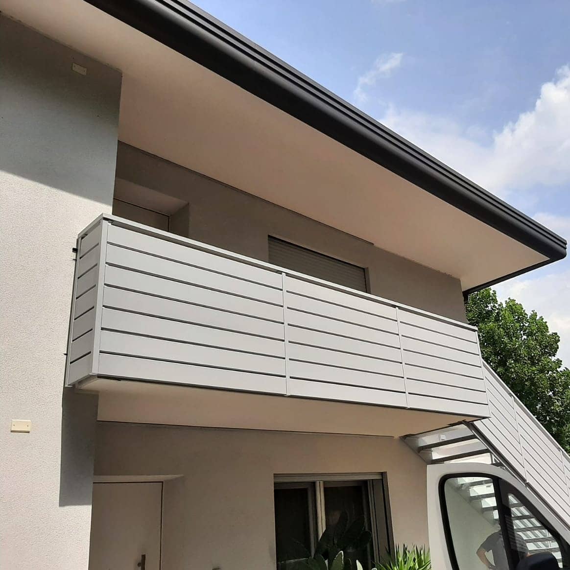 Edificio moderno con un balcone grigio e una grondaia nera sotto un cielo azzurro.