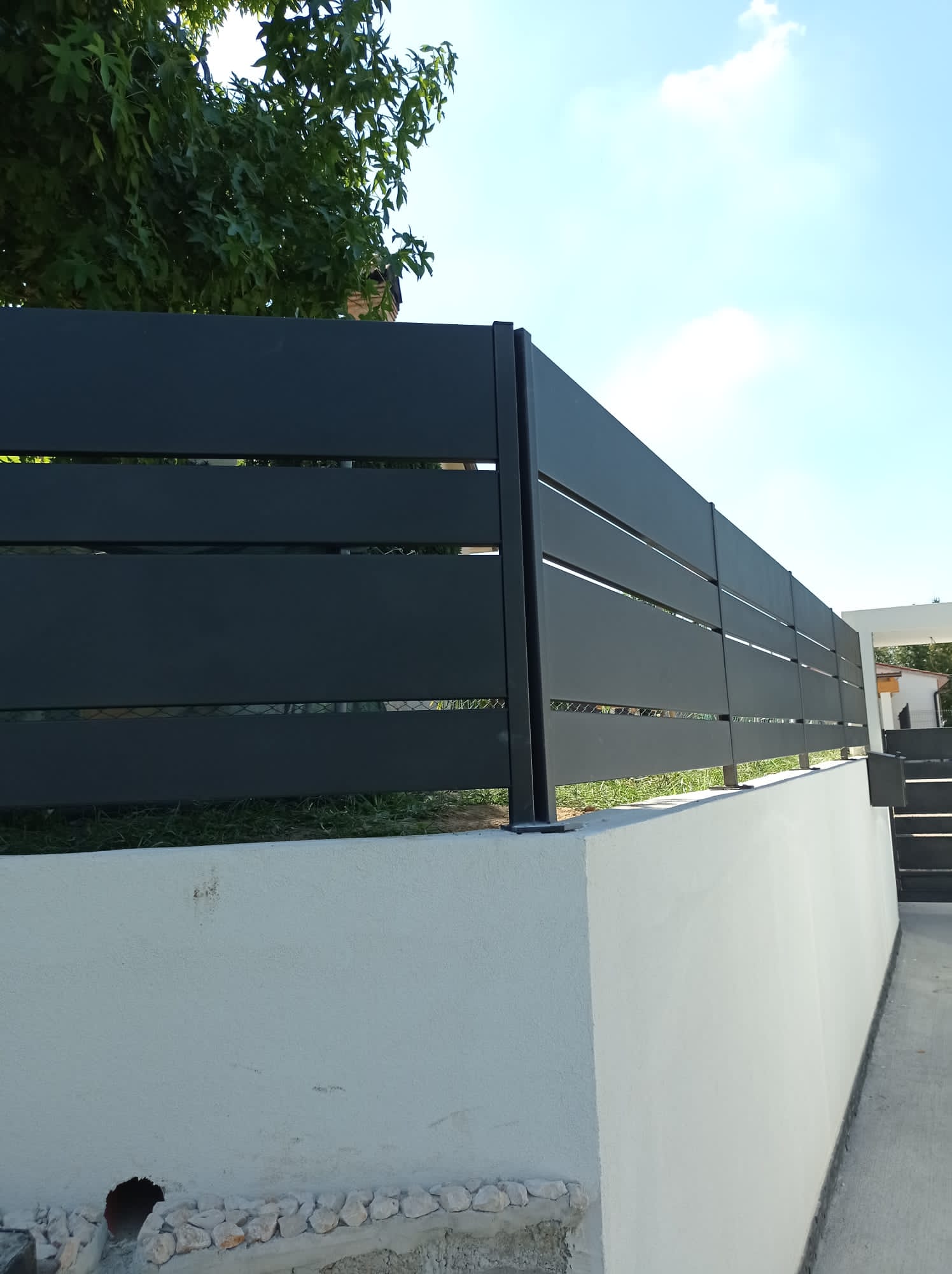 Recinzione nera a doghe orizzontali in cima a un muro di contenimento bianco. Cielo azzurro e fogliame sullo sfondo.