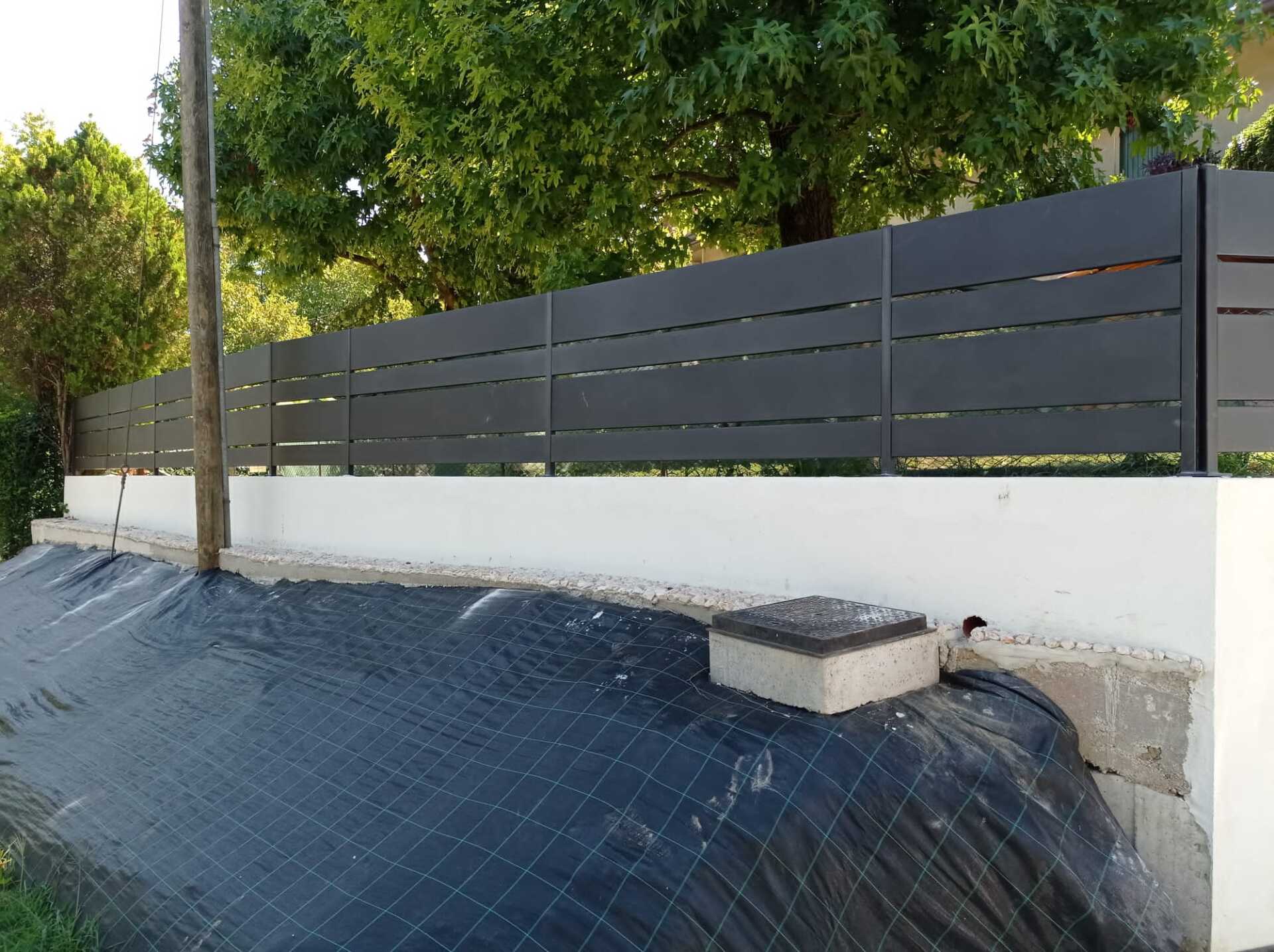 Recinzione con listelli orizzontali grigio scuro in cima a un muro bianco, di fronte a una fila di alberi.