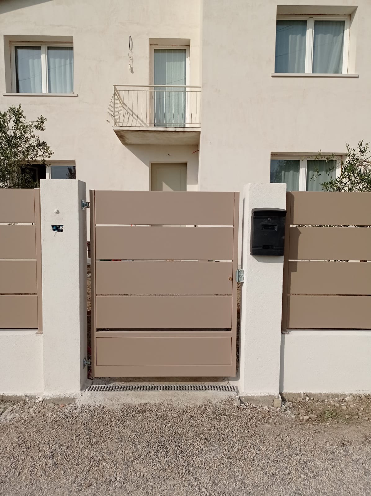 Cancello e recinzione beige di fronte a un edificio chiaro con finestre e balcone.